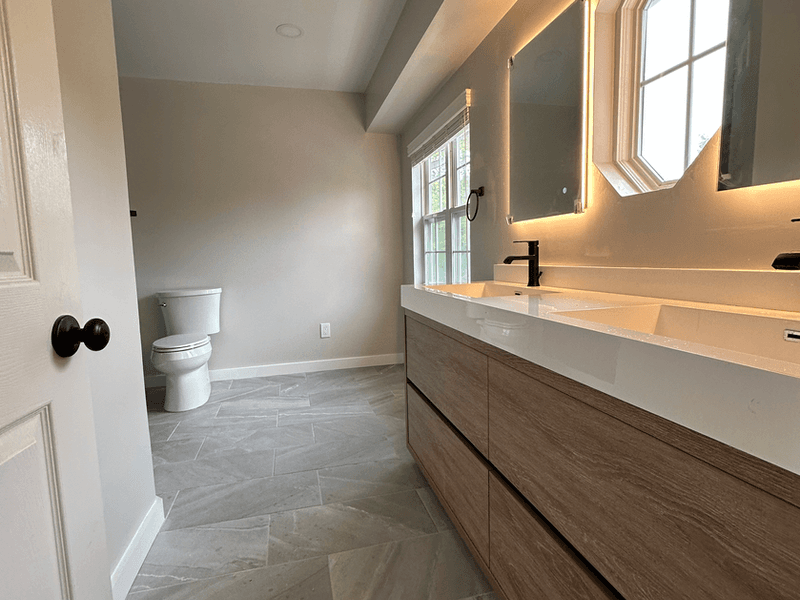 Completed modern bathroom remodel in Bound Brook, New Jersey featuring a floating wood-grain double vanity with white countertop, LED backlit mirrors, matte black faucets, and gray tile flooring