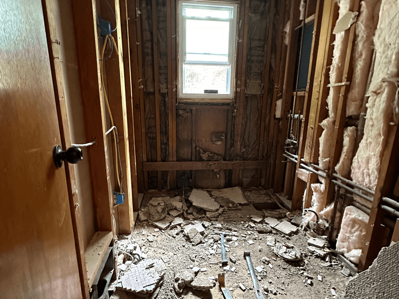 Bathroom demolition phase of a full gut renovation in Bernardsville, New Jersey showing stripped walls down to studs with exposed insulation, debris on floor, and plumbing rough-ins visible