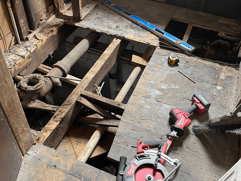 Subfloor repair during bathroom remodel in Branchburg, New Jersey with exposed floor joists, old cast iron plumbing pipes, power tools including a Milwaukee circular saw and level for precise work