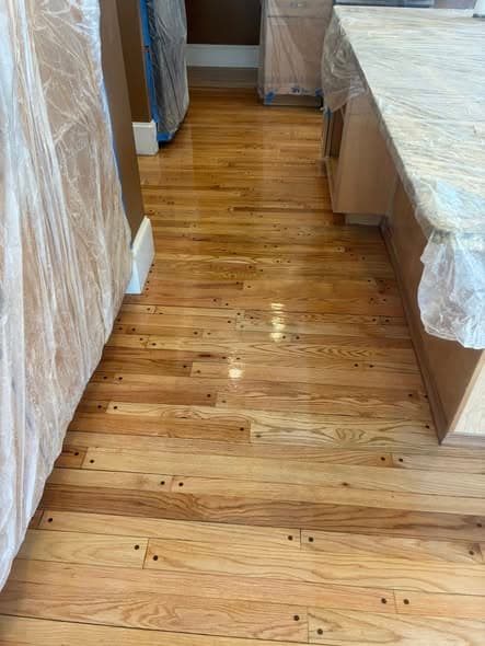 Freshly refinished natural oak hardwood floors with a glossy polyurethane finish in a kitchen hallway, furniture covered with plastic sheeting for protection during flooring installation in Somerville, New Jersey