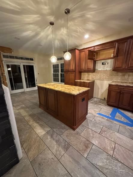 Kitchen remodeling project in progress by a general contractor in New Jersey featuring rich cherry wood cabinetry, granite countertop island with pendant lighting, tile flooring, and French patio doors in a Bridgewater area home renovation