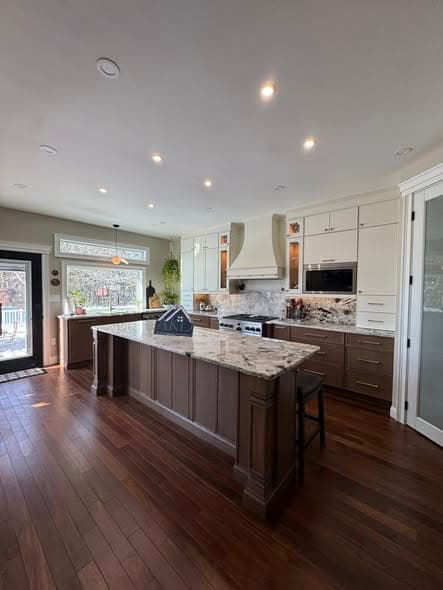 Completed luxury kitchen renovation by a New Jersey general contractor with two-tone cabinetry, white uppers and brown lowers, marble countertop island, custom range hood, hardwood flooring, and recessed lighting in a Hillsborough home remodeling project