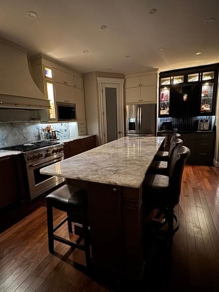 Elegant evening view of a completed kitchen remodel in New Jersey with marble countertop island, leather bar stools, professional gas range, custom range hood, illuminated display cabinet, and hardwood floors by a licensed general contractor