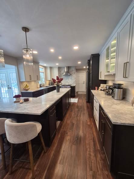 Spacious modern kitchen remodeling project completed by a New Jersey general contractor with white upper cabinets, dark navy lower cabinets, marble countertops, crystal pendant chandelier, stainless steel hood, and wide-plank hardwood floors in a Franklin Township home renovation