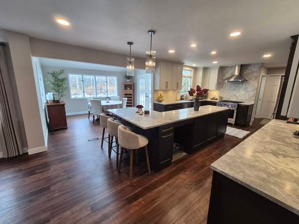 Open-plan kitchen and dining renovation by a general contractor featuring a large center island with upholstered bar stools, dark base cabinets with marble countertops, crystal pendant lighting, stainless steel range hood, and hardwood flooring in a Raritan area New Jersey home remodeling