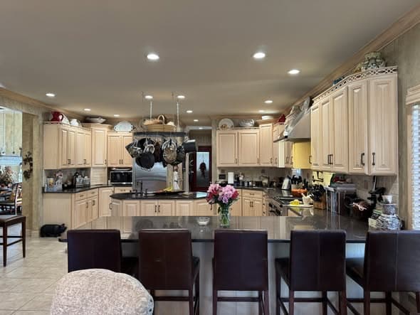 Spacious open-concept kitchen renovation in Manville, New Jersey with light maple raised-panel cabinets, dark granite countertops, a large island with brown leather bar stools, hanging pot rack, recessed lighting, stainless steel appliances, and beige tile flooring flowing into an adjacent living area
