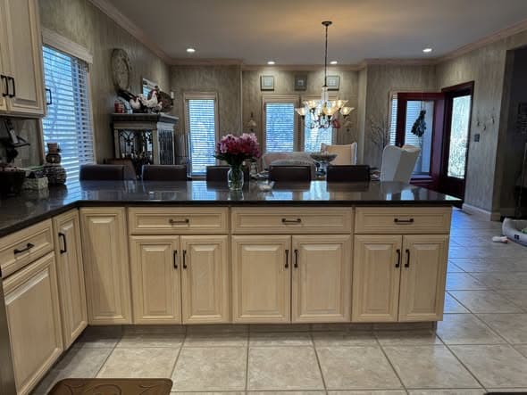 Kitchen island close-up in a Green Brook, New Jersey home renovation showing light maple cabinetry with dark bronze hardware, polished black granite countertops, beige ceramic tile flooring, and a view through to the dining room with a chandelier and natural light from multiple windows