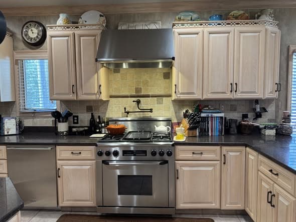 Kitchen renovation cooking area in Montgomery, New Jersey featuring light glazed maple cabinets with decorative crown molding, a professional-style stainless steel gas range with large stainless hood, pot filler faucet, stone tile backsplash, dark granite countertops, and display shelving above the upper cabinets