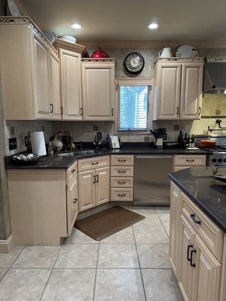 Corner kitchen view of a Bridgewater, New Jersey kitchen remodel with light glazed cabinets, dark granite countertops, stainless steel dishwasher, stone tile backsplash, recessed lighting, a window above the sink area with blinds, ceramic tile flooring, and a comfort mat in front of the sink