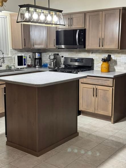 Completed kitchen renovation in Somerville, New Jersey with warm brown stained shaker cabinets, light gray speckled quartz countertops, a center island with matching cabinetry, stone ledger backsplash, black stainless microwave and gas cooktop, modern geometric pendant light fixture, and large-format beige tile flooring