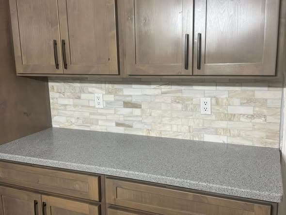 Close-up detail of a kitchen remodel backsplash and countertop in Bound Brook, New Jersey showing brown stained shaker-style upper cabinets with matte black bar pulls, a natural stone linear mosaic tile backsplash in cream and tan tones, and gray speckled quartz countertop with electrical outlets