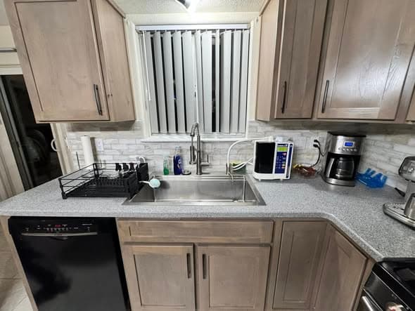 Kitchen sink area in a Hillsborough, New Jersey renovation featuring light brown stained cabinets, gray quartz countertops, a stainless steel single-bowl sink with pull-down faucet, stone mosaic tile backsplash, a window with vertical blinds, black dishwasher, dish drying rack, microwave, and coffee maker
