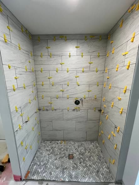 Shower tile installation in progress with gray marble-look porcelain wall tiles and yellow leveling clips ensuring even spacing, herringbone mosaic marble floor tile visible below, bathroom tile work in Bound Brook, New Jersey