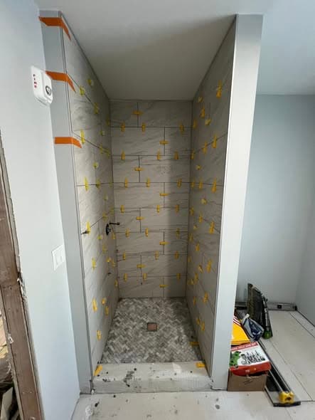 Walk-in shower tile installation in progress showing gray marble-look porcelain wall tiles with yellow and orange leveling spacers, herringbone pattern mosaic shower floor tile, bathroom renovation in Warren Township, New Jersey