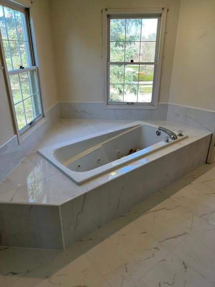 Bathroom tile installation featuring white Calacatta marble-look porcelain tile on bathtub surround and floor, corner jetted soaking tub with polished tile deck, elegant master bathroom renovation in Hillsborough, New Jersey